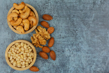 Close-up view from above of wooden bowls with pine nuts and cashews, which stand on the side surrounded by walnuts and almonds on a gray background with space for text. The concept of healthy snacks