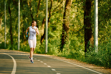 Fototapeta premium Sporty woman running along a serene path surrounded by lush greenery in bright daylight