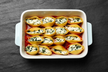 Delicious conchiglie pasta with ricotta cheese, spinach and tomato sauce in baking dish on black textured table, top view