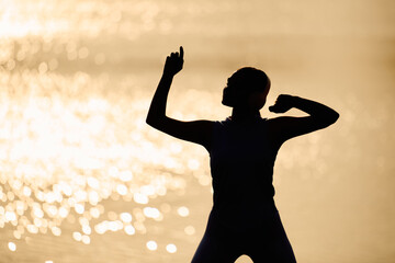 Fototapeta premium Young beautiful sporty woman expressing joy near shimmering water at sunset