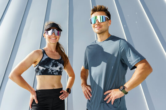 Couple poses confidently in athletic wear with stylish sunglasses against modern building backdrop