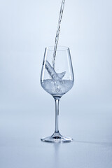 Clear stemmed glass being filled with transparent water, showing splash and bubbles, against blue background. Water being poured into glass. Mock up. Concept of nutrition, hydration, drink.