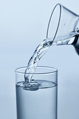 Close-up photo of water being poured from pitcher into clear glass, showing smooth stream and ripples, against light background. Mock up. Concept of nutrition and hydration, drink