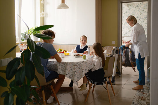 A family gathers around the breakfast table as children prepare for school. The atmosphere is warm, with the mother organizing a backpack and the children eating.