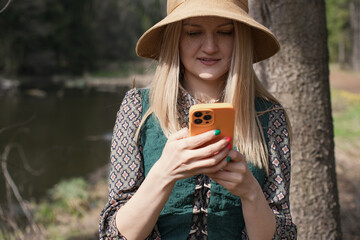 A young blonde woman in a straw hat and vintage dress is standing in a serene forest by the river,...