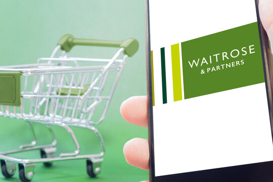 Mini supermarket trolley with green handle next to Waitrose And Parners logo on screen on green background. United Kingdom supermarket chain