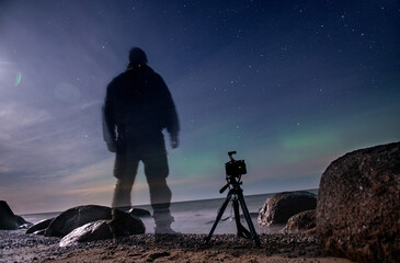 Fototapeta premium Traveler shooting northern lights on the seashore under a starry night sky with a tripod.