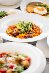 A delicious plate of penne pasta topped with meatballs and fresh herbs, garnished with microgreens. Surrounding dishes include salads and a fried egg, creating an inviting dining scene.