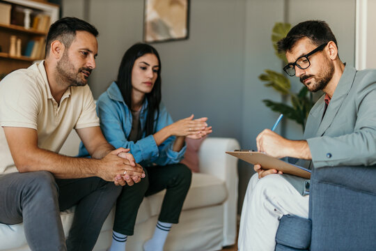 Worried couple explaining problems to psychologist taking notes during therapy session