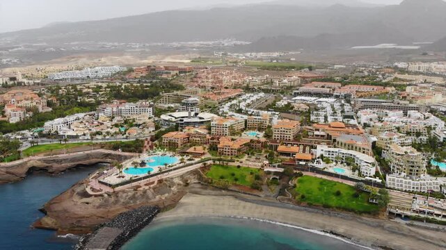 Aerial view of Costa Adeje city, Tenerife, Canary islands. 4K video with ocean coast, beach, ocean, city villas, resorts, quay, mountains. Aerial push-in shot. Footage during sand storm calima