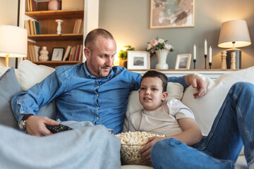 Home cinema. Happy father and son watching film on TV and eating popcorn, resting on comfy sofa in...