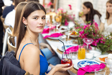 A young charming girl in a blue dress is sitting at a festive table and holding a glass. A cute teenager in an elegant suit drinks a cocktail at a festive dinner.