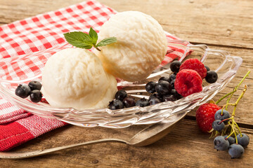 Two scoops of vanilla ice cream with raspberries and blueberries in glass bowl on rustic table