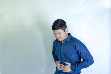 Young Asian man in blue shirt holding stomach with both hands, his face showing pain expression, stomachache with white studio background. Disease concept