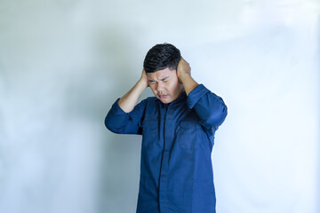 young asian man in blue shirt holding head with hands, his face showing pain expression, headache with white studio background. Concept of disease, treatment, health care