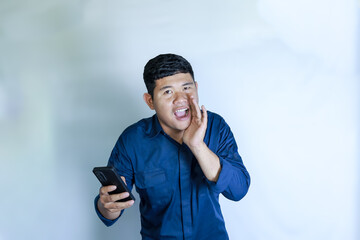 A young man feels happy, shouting loudly with his hands at the side of his mouth and holding a smartphone
