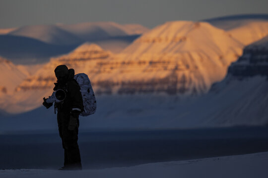 Silhouetted photographer on icy terrain in Elveneset, Svalbard, with a snow-covered mountain at sunset.
