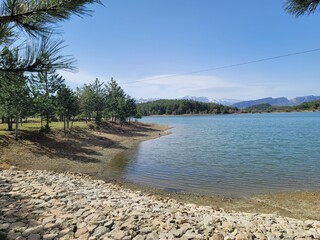 Lakeside scene with a tree-lined shore, tranquil blue waters, and rocky edges stretching into the horizon. A pristine natural escape with lush greenery and a peaceful atmosphere.