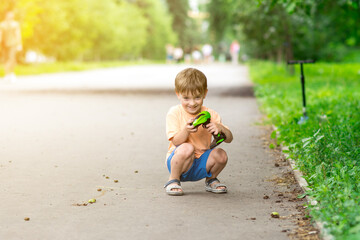 A happy young boy squats on a park path, holding a green remote control, enjoying his toy car. The sunlight and greenery create a warm outdoor scene.