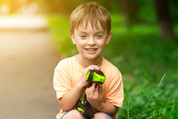 A cheerful boy with light hair plays with a green remote-control car in a sunny park. The vibrant nature and golden light highlight a carefree childhood moment, great for outdoor fun and kids' leisure