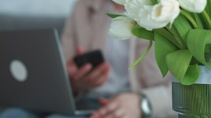 A vase of flowers on the table and a man who is clicking a phone in his hands.
A man in a cozy bright room sits on a sofa and clicks a phone