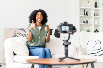 An enthusiastic African-American woman gives a thumbs up while recording a motivational vlog, her...