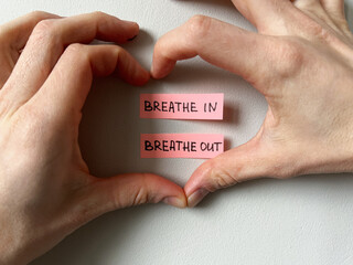 Two hands form a heart shape around pink sticky notes with the words “Breathe In” and “Breathe Out,” symbolizing mindfulness, meditation and conscious breathing 