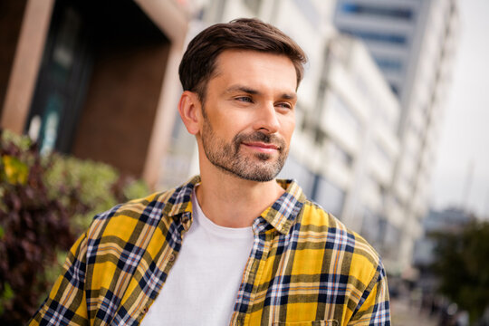 Portrait of a young man outdoors in a casual yellow checkered shirt, standing confidently with an urban backdrop and a thoughtful smile