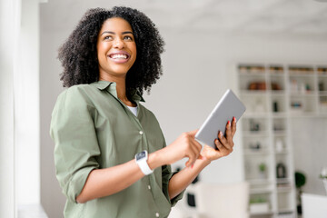African-American woman is confidently handling a tablet, standing in a well-lit room with a smile. Cheerful female student or remote worker using digital tablet