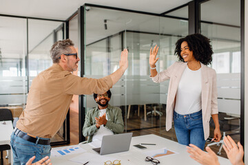 A team leader and a businesswoman exchange a high-five in a collaborative office setting while teammates cheer. A representation of teamwork, celebration, and motivation in the corporate world.