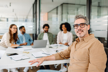 Mature businessman in glasses and brown shirt confidently seated in a modern office with a diverse team engaged in a meeting in the background.