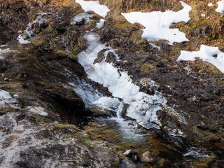 Melting snow creates small cascading waterfall over dark mountain rocks in early spring
