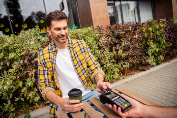 Young man outdoors in checkered shirt holding coffee and paying with a card on a portable terminal,...