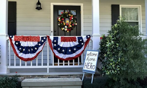 Patriotic Porch Decor Bunting And Floral Wreath Displayed On Porch Railing With Sign And Bushes On Gray House Exterior Decorated For Holidays Including Fourth of July Celebration And Independence Day