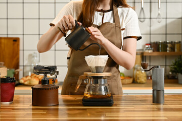 A woman is making coffee and pouring it into a cup. The scene is set in a kitchen with various items such as a scale, a cup, a bottle, and a bowl. The woman is wearing an apron.
