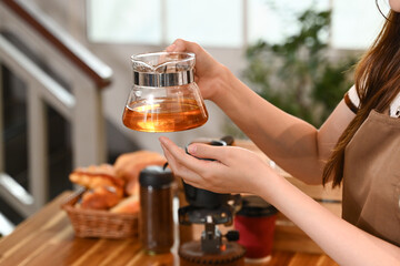A woman is making tea in a kitchen. She is wearing an apron and holding a tea strainer.