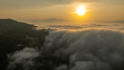 Breathtaking aerial view of the sun rising over a mountain valley covered in a dense sea of rolling clouds. The golden light illuminates the mist, creating a dramatic and ethereal atmosphere at dawn