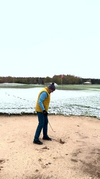 A golfer in a yellow vest hits a bunker shot on a snow-covered golf course, striking a blue ball towards the green near the flag.
