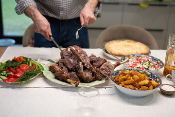 Hands carving a whole roasted goose on a platter, surrounded by side dishes like roasted potatoes, fresh salad, and a pie, creating a festive dining atmosphere during New Year celebration evening