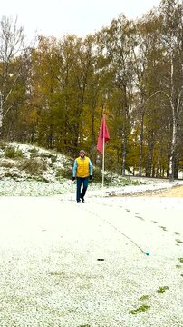 A golfer in a yellow vest and winter hat is putting on a snow-covered green near a red flag, surrounded by autumn trees.
