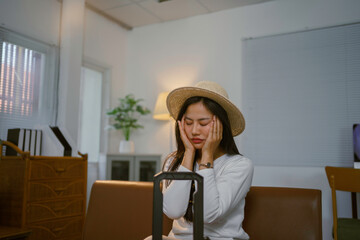 Asian woman sitting on sofa with suitcase and holding head in hands, suffering from headache or...