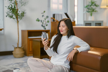 Young woman relaxing cross legged near sofa, scrolling smartphone and messaging friends in cozy living space