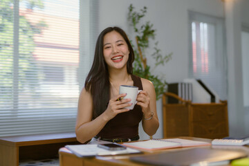 Young Asian businesswoman smiling and enjoying a warm cup of coffee during a refreshing work break in her modern office space