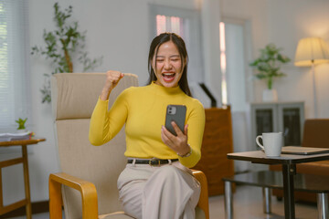 Young Asian businesswoman raising fist while looking at mobile phone, celebrating business success in home office