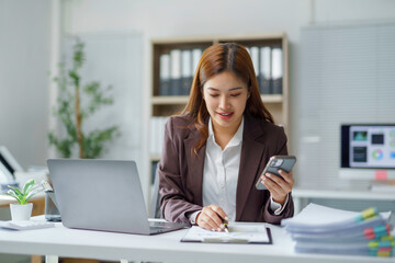 Young businesswoman comparing information on smartphone and financial documents while working at office desk
