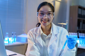 Fototapeta premium Female scientist in white lab coat examining blue liquid test tube, smiling confidently during medical research experiment