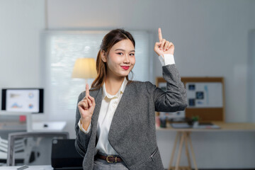 Portrait of a smiling asian businesswoman pointing up with index fingers while standing in a modern office