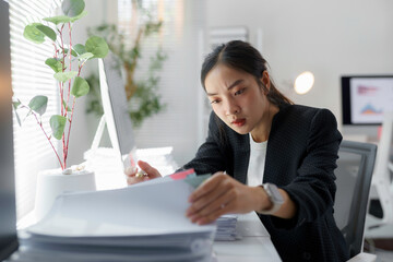 Young professional managing paperwork at workplace, focused on finding specific information among...