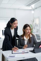 Two asian businesswomen analyzing financial charts and graphs using tablet computer in office, discussing company growth and financial performance