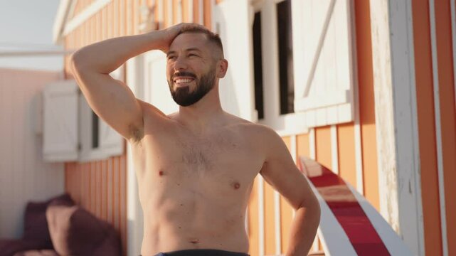 A shirtless man with a beard smiles at the beach as he runs his hand through his hair. A surfboard leans against a nearby beach hut.